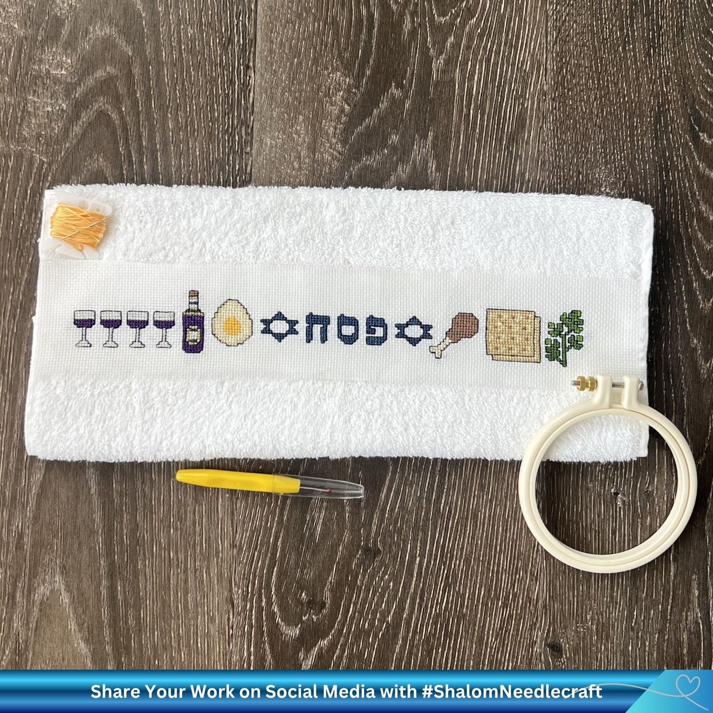 Embroidery hoop with cross-stitched Pesach design on a towel, surrounded by embroidery tools on a wooden surface.