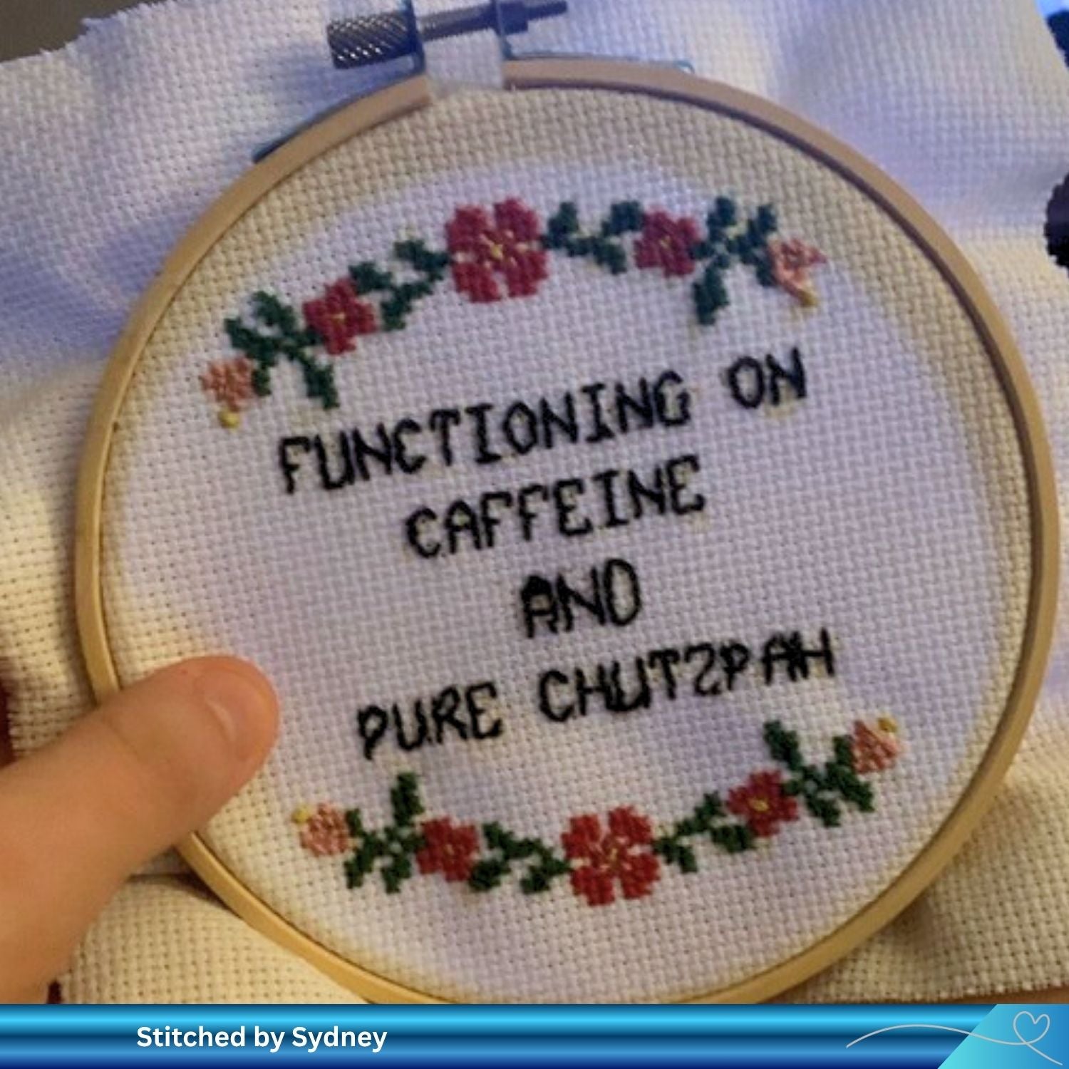 Hand holding a finished cross-stitched hoop with text 'Functioning on caffeine and pure chutzpah' on Aida