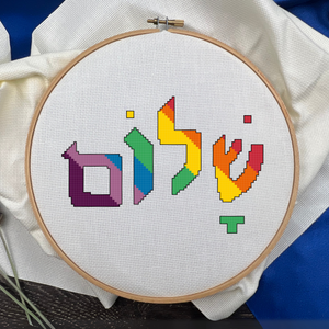 Embroidery hoop with colorful Hebrew letters cross stitched onto a white aida fabric background