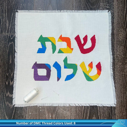 Rainbow colored Shabbat Shalom cross stitch design on challah cover against a wooden background