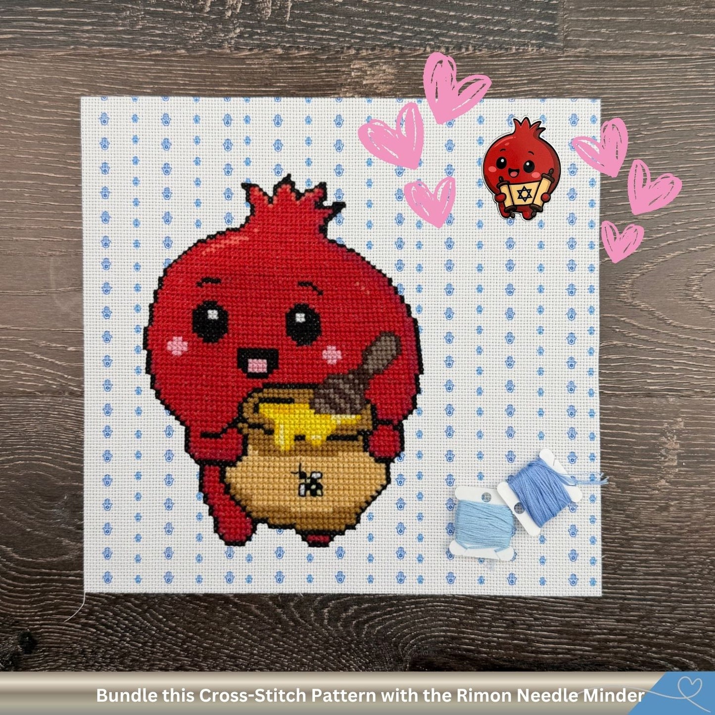 A cross-stitch pattern preview showing a cute red pomegranate character holding a yellow honey jar, stitched on white Aida cloth with light blue Hamsa hands. Pink hearts encircle a matching RImon pomegranate needle minder. The text banner at the bottom reads 'Bundle this Cross-Stitch Pattern with the Matching Needle Minder'.