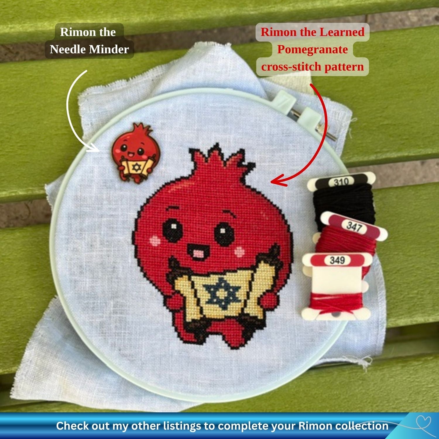 A completed cross-stitch project in a an embroidery hoop, featuring a red pomegranate character holding a gold scroll with a Star of David. The hoop rests on a green wooden surface, next to thread spools and two text callouts reading 'Rimon the Needle Minder' and 'Rimon the Learned Pomegranate cross-stitch pattern'.