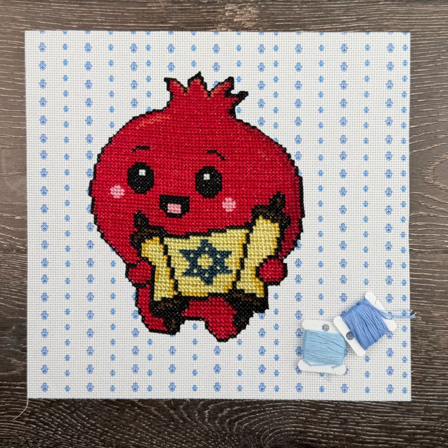 Cross-stitched design of a red Pomegranate character holding a Torah with blue thread on a white fabric background.