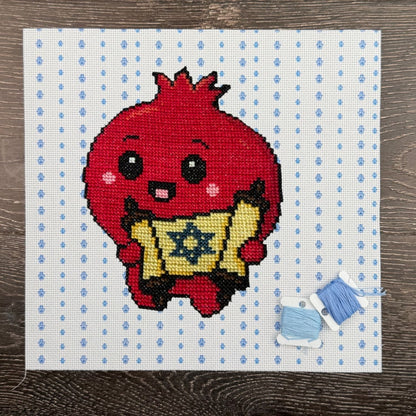 Cross-stitched design of a red Pomegranate character holding a Torah with blue thread on a white fabric background.