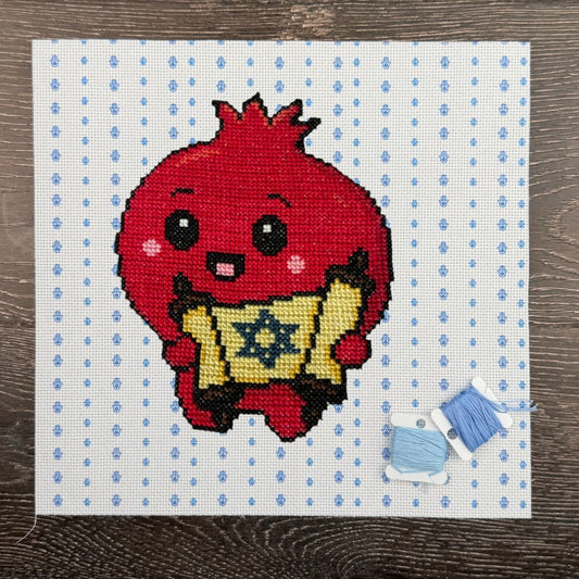 Cross-stitched design of a red Pomegranate character holding a Torah with blue thread on a white fabric background.