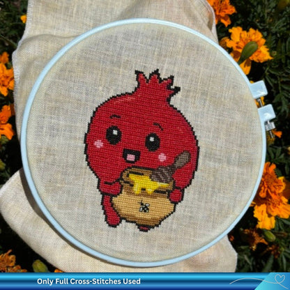 A completed cross-stitch embroidery in a white hoop, featuring a cute red pomegranate character holding a pot of honey, set against a beige linen fabric with orange flowers in the background.