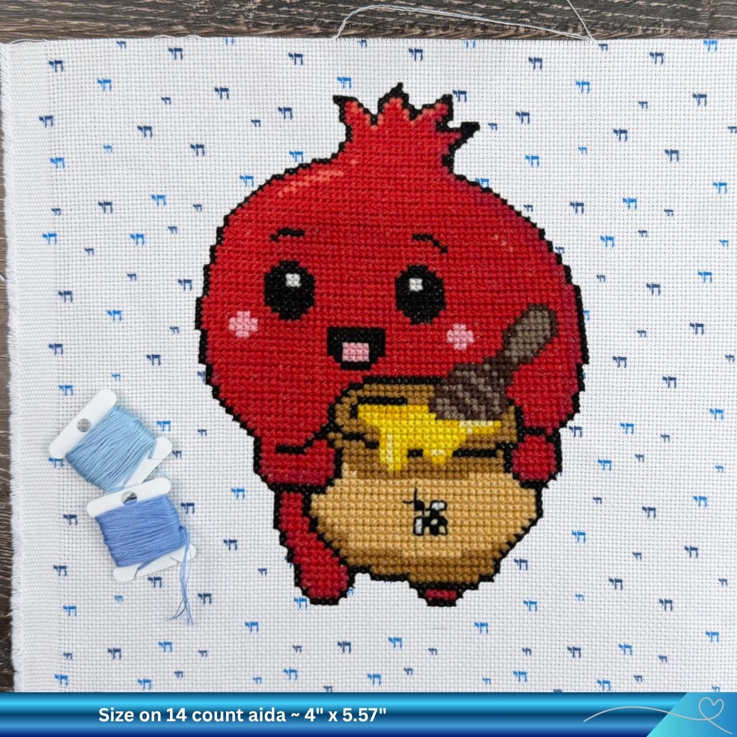 A completed cross-stitch design of a cute red pomegranate holding a pot of honey, stitched on white Aida fabric with a blue Chai pattern. Two spools of thread are placed on the left side, and a the size of the finished product on 14 count aida is in a text banner is at the bottom.