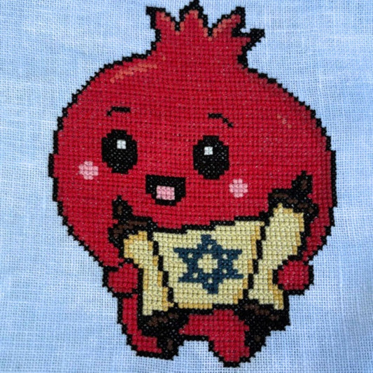 A close-up of a cross-stitch or needlepoint design on light blue fabric, featuring a cute red pomegranate character holding a beige scroll with a blue Star of David.