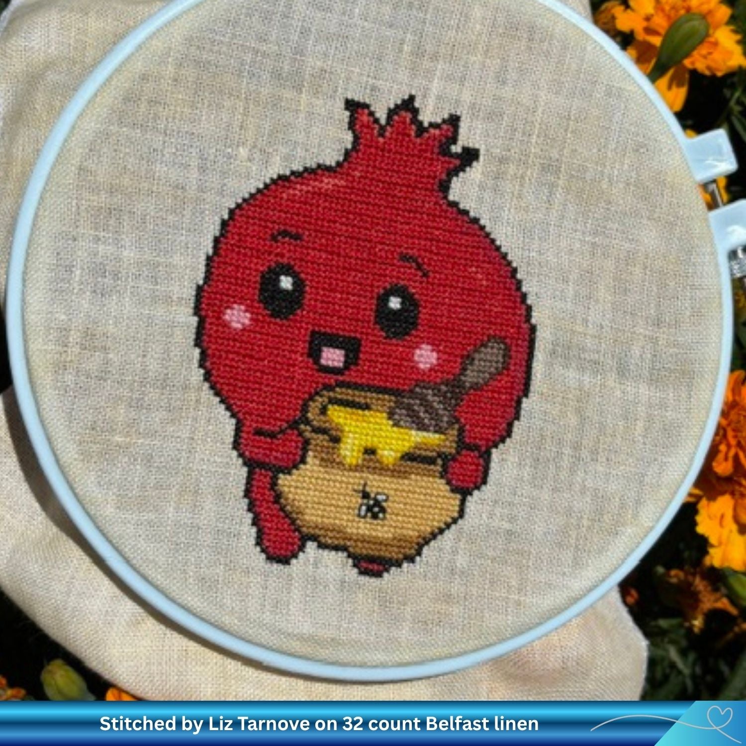 A close-up of a completed cross-stitch design on beige Belfast linen, showing a red cartoon pomegranate holding a yellow honey pot. The embroidery is held in a white hoop, with orange flowers blurred in the background.