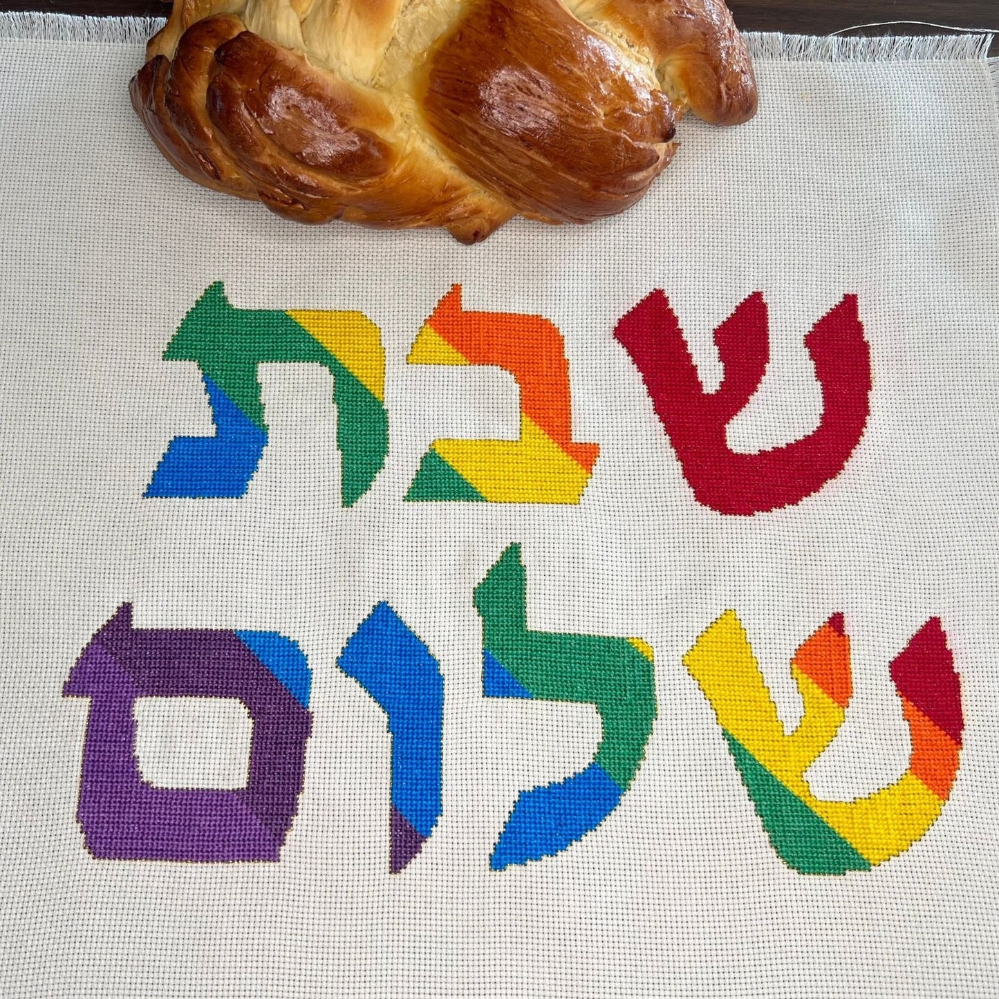 Cross-stitched rainbow Hebrew letters on fabric with a loaf of Challah bread