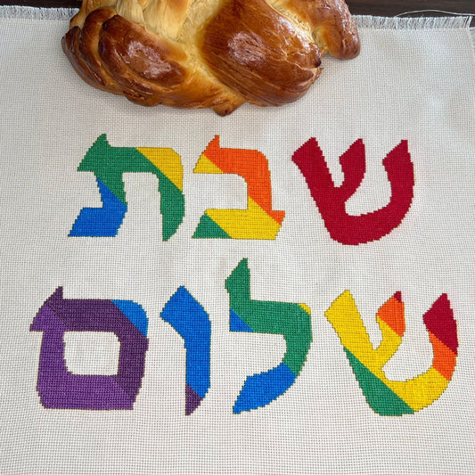 Cross-stitched rainbow Hebrew letters on fabric with a loaf of Challah bread