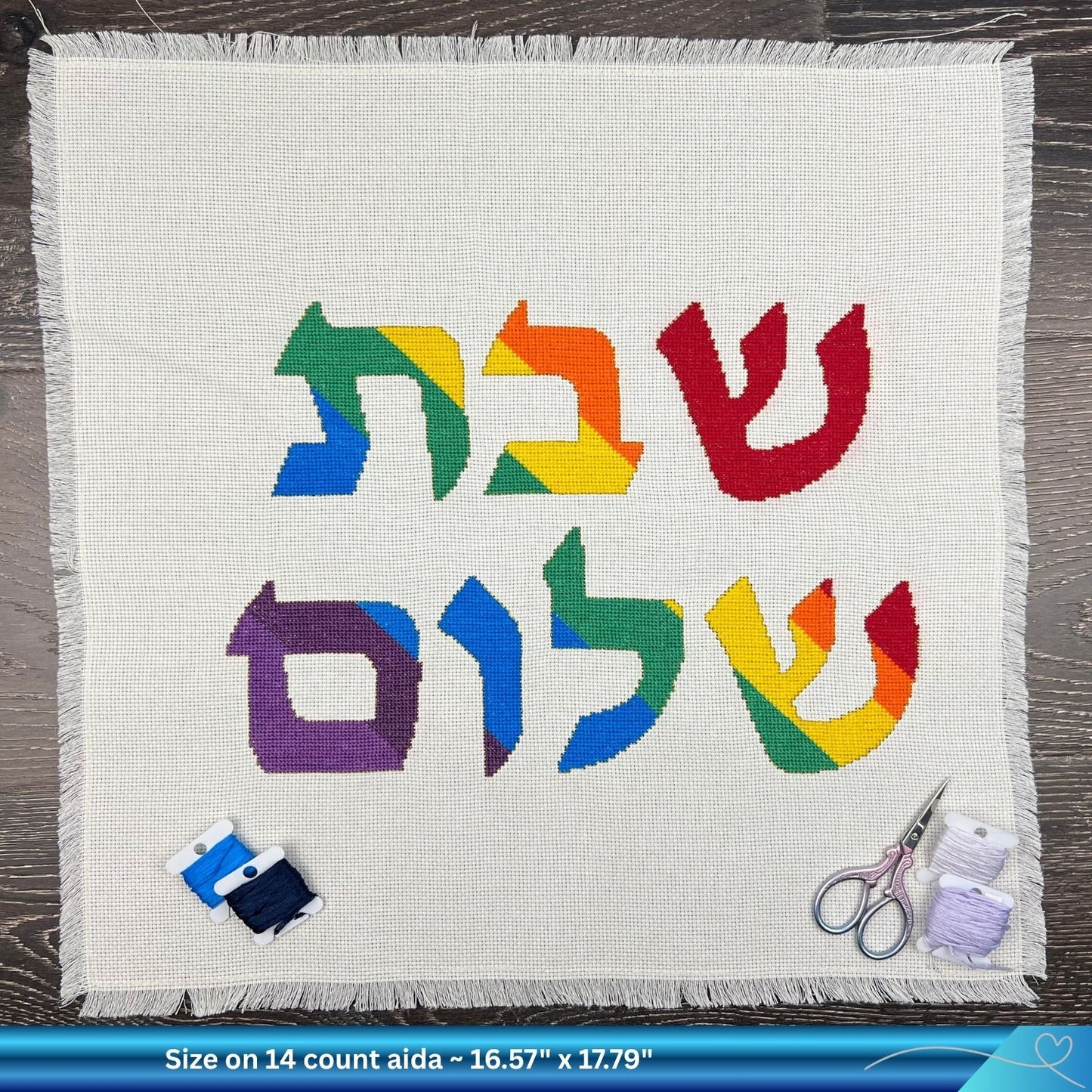 Colorful Hebrew text saying Shabbat Shalom on a white Aida cotton with embroidery thread and scissors on a wooden surface