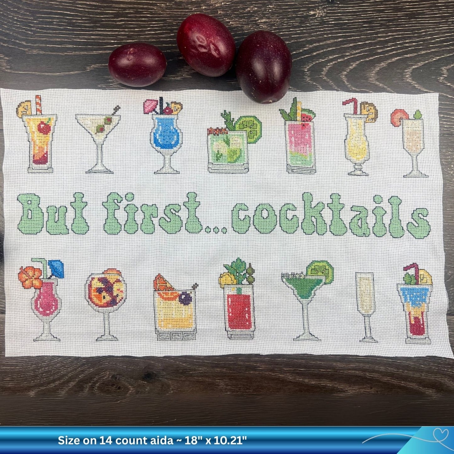 Piece of white Aida fabric with the cross-stitched design of cocktail glasses and 'But first... cocktails' text on a wooden surface. There are some summer passion fruit towards the top of the image.