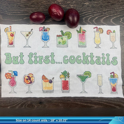 Piece of white Aida fabric with the cross-stitched design of cocktail glasses and 'But first... cocktails' text on a wooden surface. There are some summer passion fruit towards the top of the image.