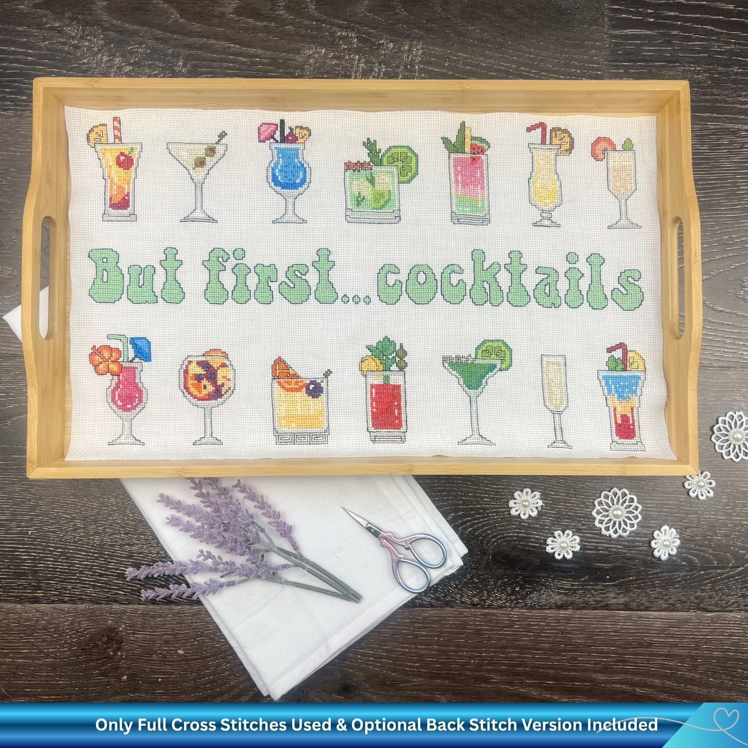 Cross-stitched pattern with cocktail designs and 'But first... cocktails' text on a wooden tray. There are also embroidery items and some lavender around the tray for summer.