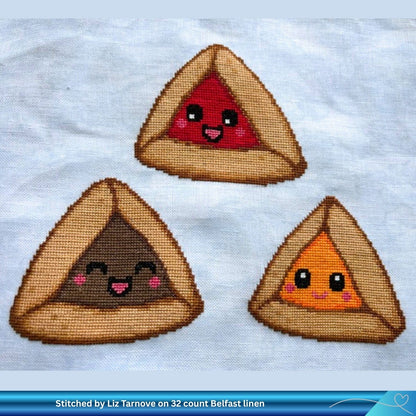 Three Hamantaschen embroidered cross-stitch designs each with a cute smiling face. One is red, one is brown, and one is orange, all on a white linen fabric background.