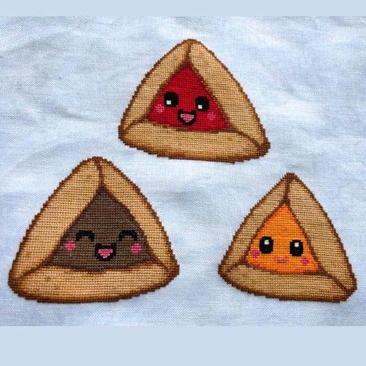Cross-stitch pattern with smiling hamantaschen on a white linen background.