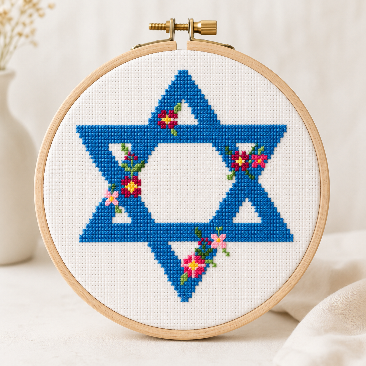 Embroidery hoop with a blue Star of David cross-stitch design on white fabric, surrounded by flowers.
